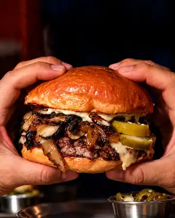 Dos manos sosteniendo una burger con hongos, cebolla caramelizada, queso y pepinillos
